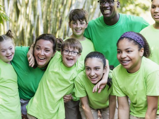 Multi-ethnic children having fun at summer camp on a field trip to a park. A camp counselor, a mid adult woman in her 30s, is standing between an 11 year old girl and 12 year old boy who both have down syndrome.  The boy is smiling at the camera.